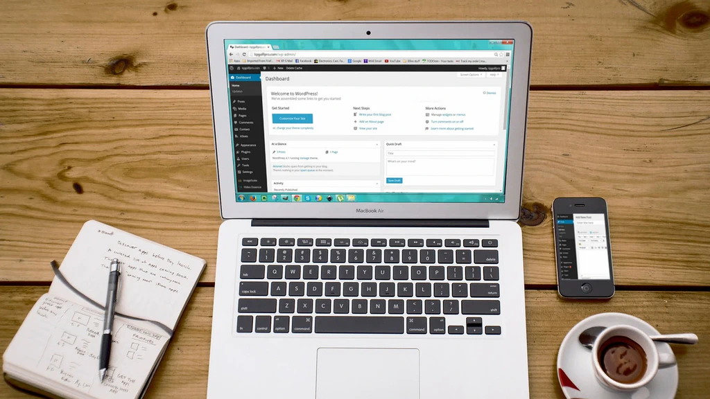 Tampilan dashboard WordPress pada layar laptop MacBook Air di atas meja kayu dengan buku catatan, ponsel, dan secangkir kopi.